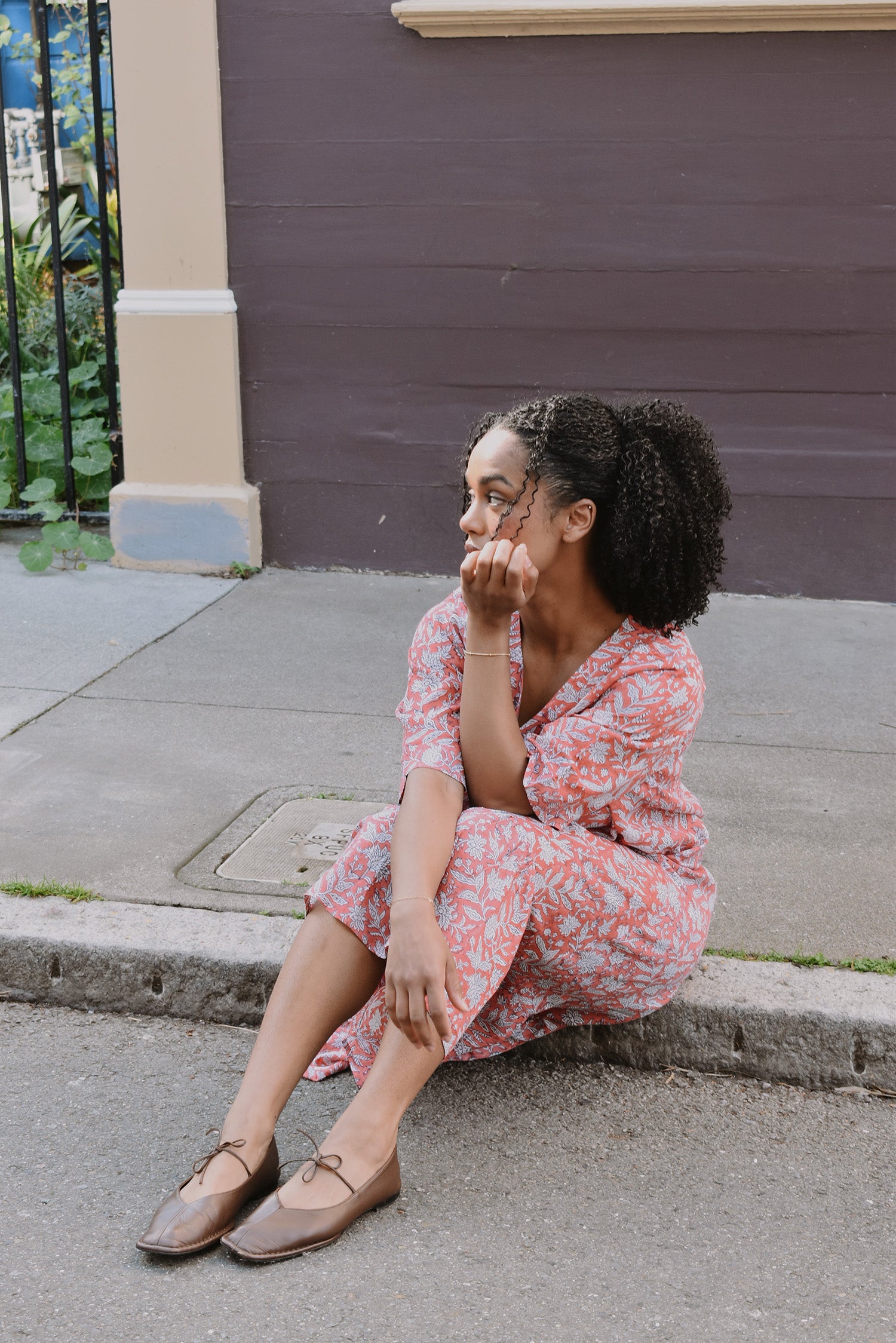Woman in a pink floral dress sitting on a sidewalk against a building.
