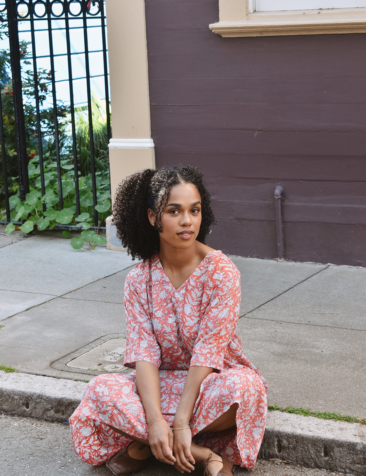 Woman in a pink floral dress sitting on a sidewalk next to a building.