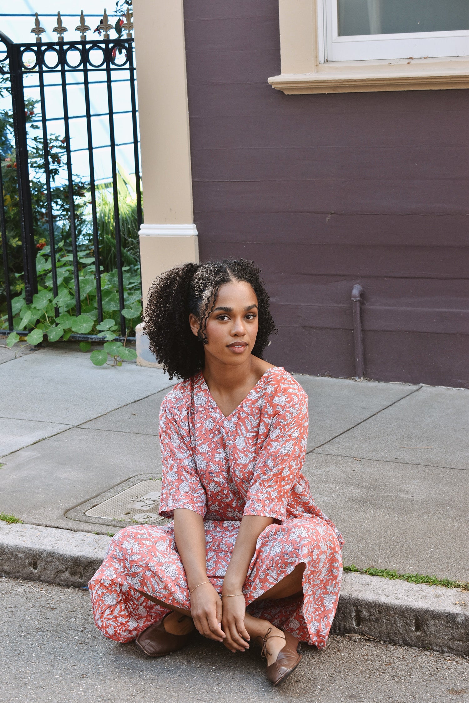 Woman in a pink floral dress sitting on a sidewalk next to a building.