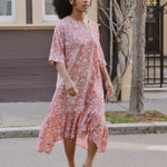 Woman in a pink floral dress standing on a street with a building and tree in the background