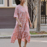 Woman in a pink floral dress standing on a street with a building and tree in the background