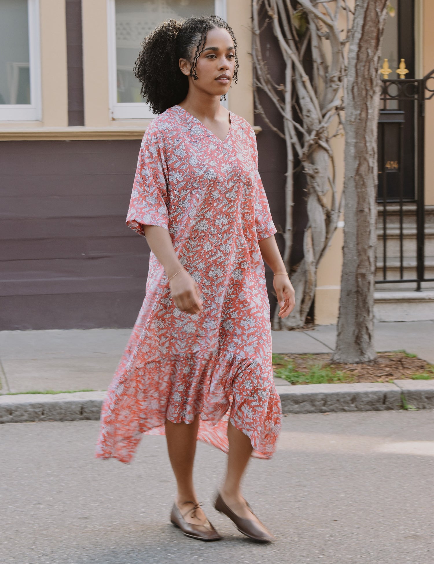 Woman in a pink floral dress standing on a street with a building and tree in the background