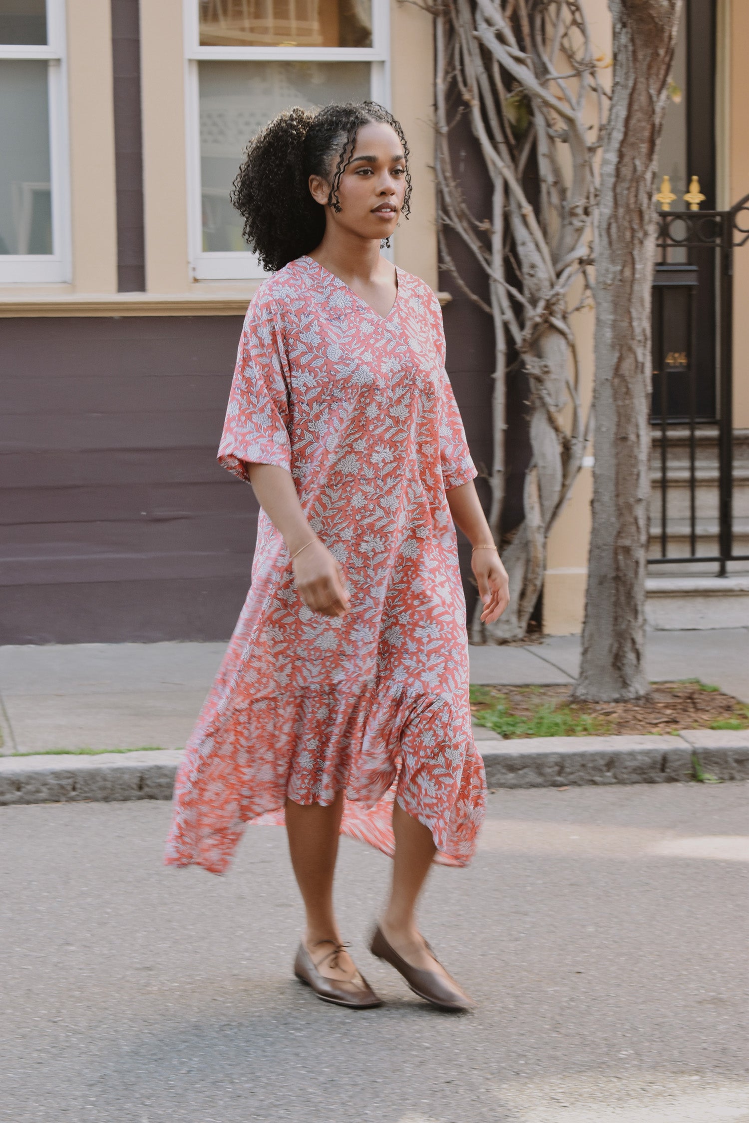 Woman in a pink floral dress standing on a street with a building and tree in the background