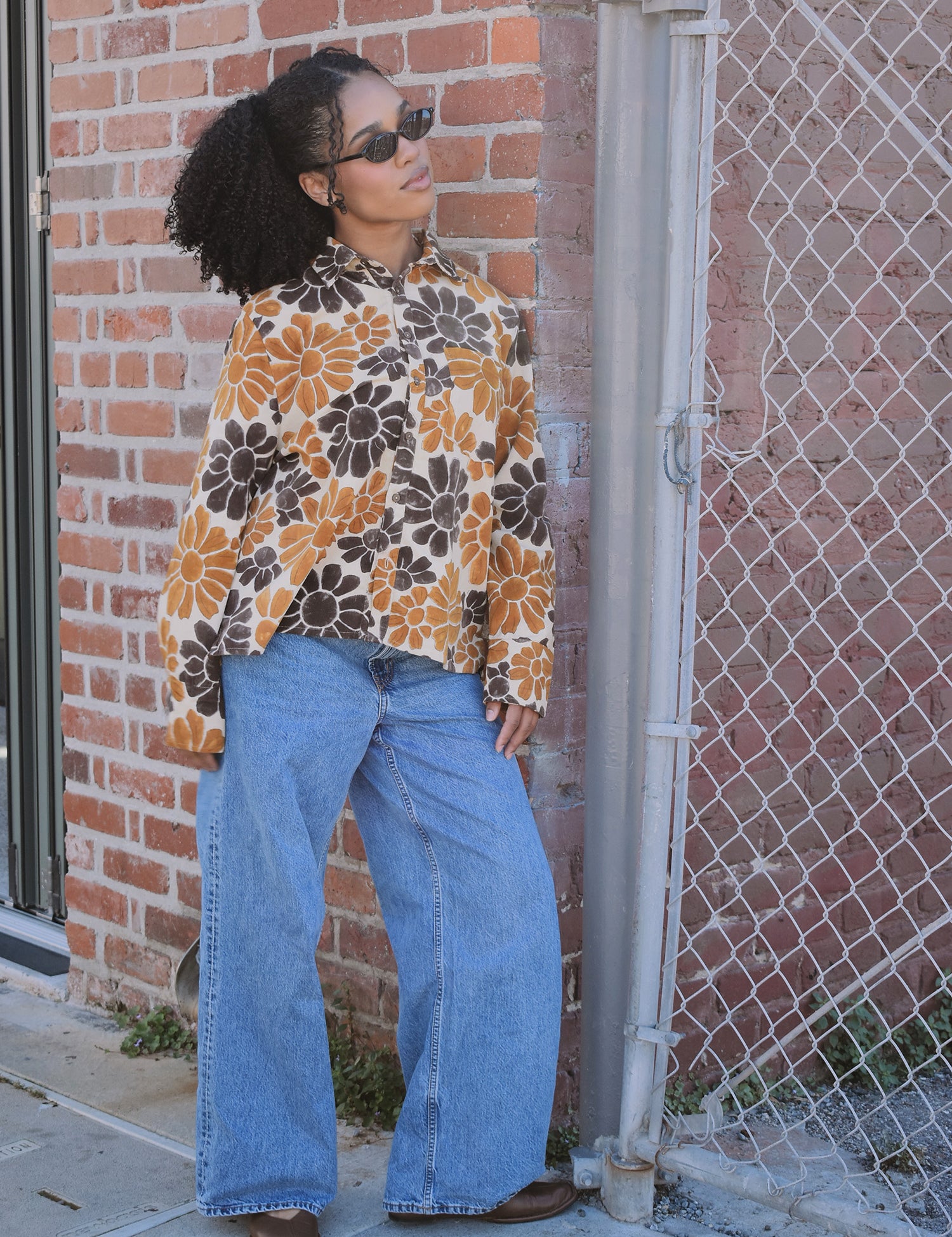 Person wearing a floral corduroy shirt and blue jeans leaning against a brick wall.
