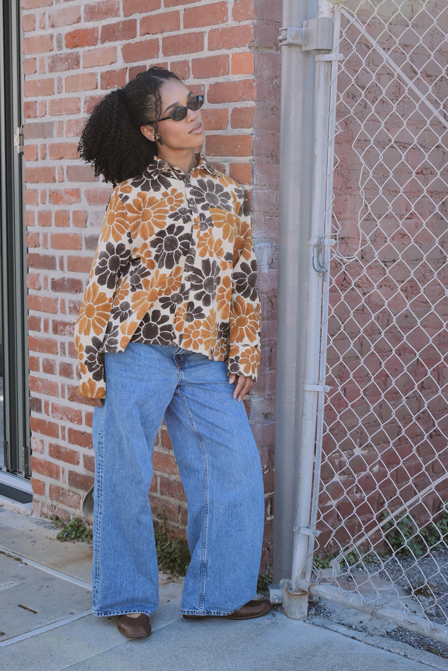 Person wearing a floral corduroy shirt and blue jeans leaning against a brick wall.