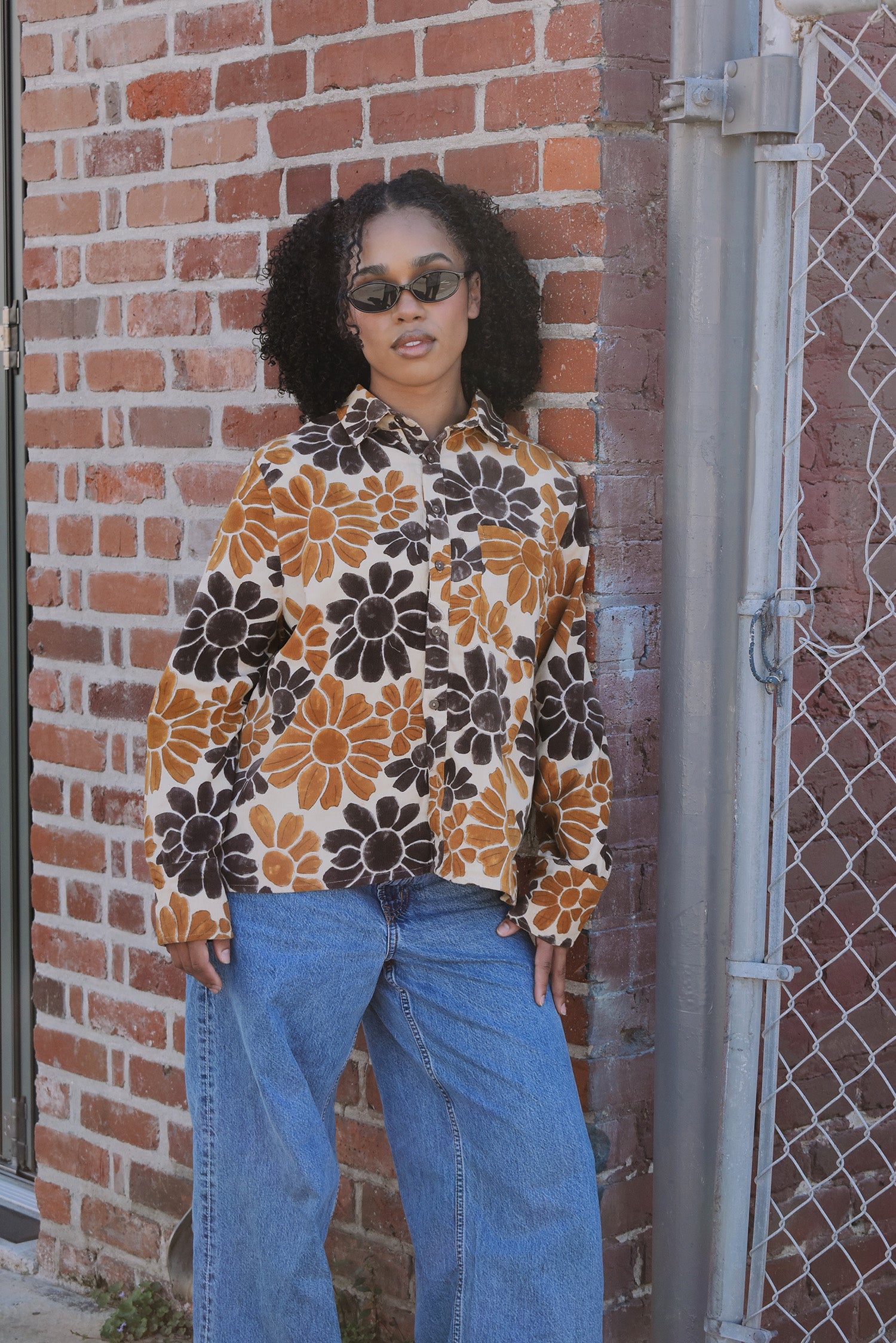 Person wearing a floral patterned corduroy shirt and blue jeans standing against a brick wall.