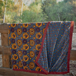 Blue velvet blanket with yellow sunflower and floral prints draped over a wooden fence, with trees in the background. One corner of blanket is draped open to show reversible print. 