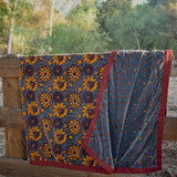 Blue velvet blanket with yellow sunflower and floral prints draped over a wooden fence, with trees in the background. One corner of blanket is draped open to show reversible print. 