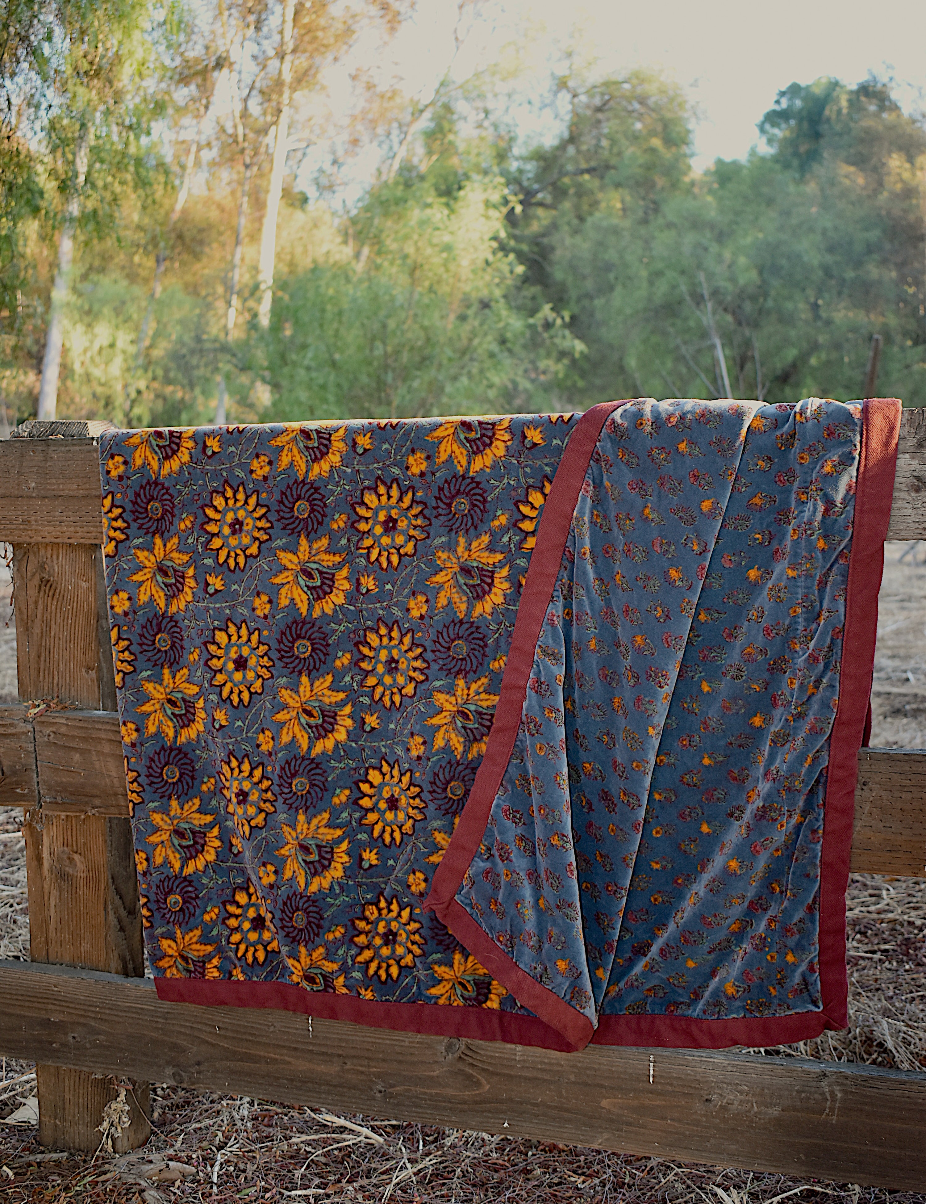 Blue velvet blanket with yellow sunflower and floral prints draped over a wooden fence, with trees in the background. One corner of blanket is draped open to show reversible print. 