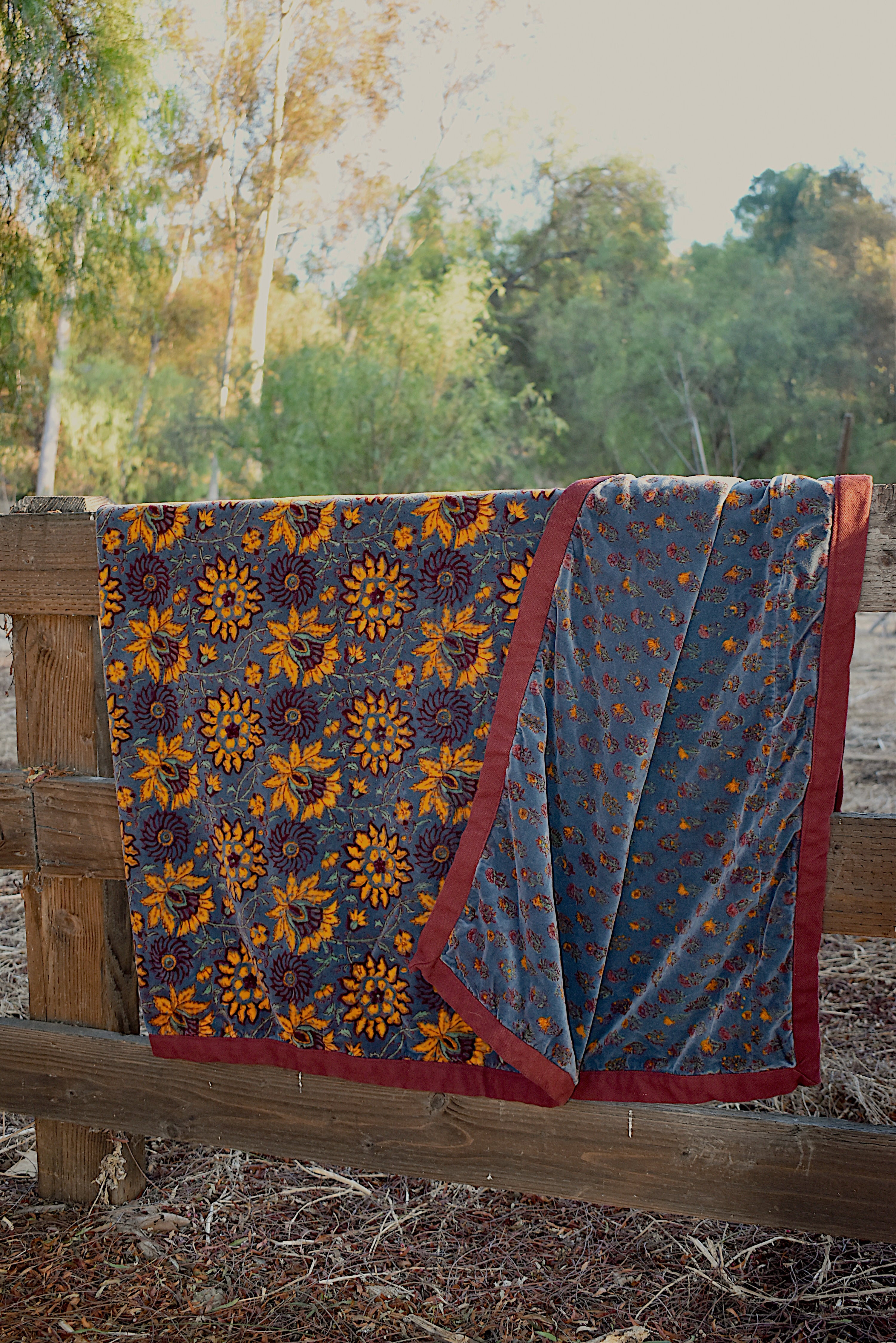 Blue velvet blanket with yellow sunflower and floral prints draped over a wooden fence, with trees in the background. One corner of blanket is draped open to show reversible print. 