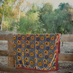 Blue velvet blanket with large yellow floral print and red cotton piping draped over a wooden fence in a rustic setting with trees in the background.