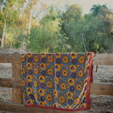 Blue velvet blanket with large yellow floral print and red cotton piping draped over a wooden fence in a rustic setting with trees in the background.