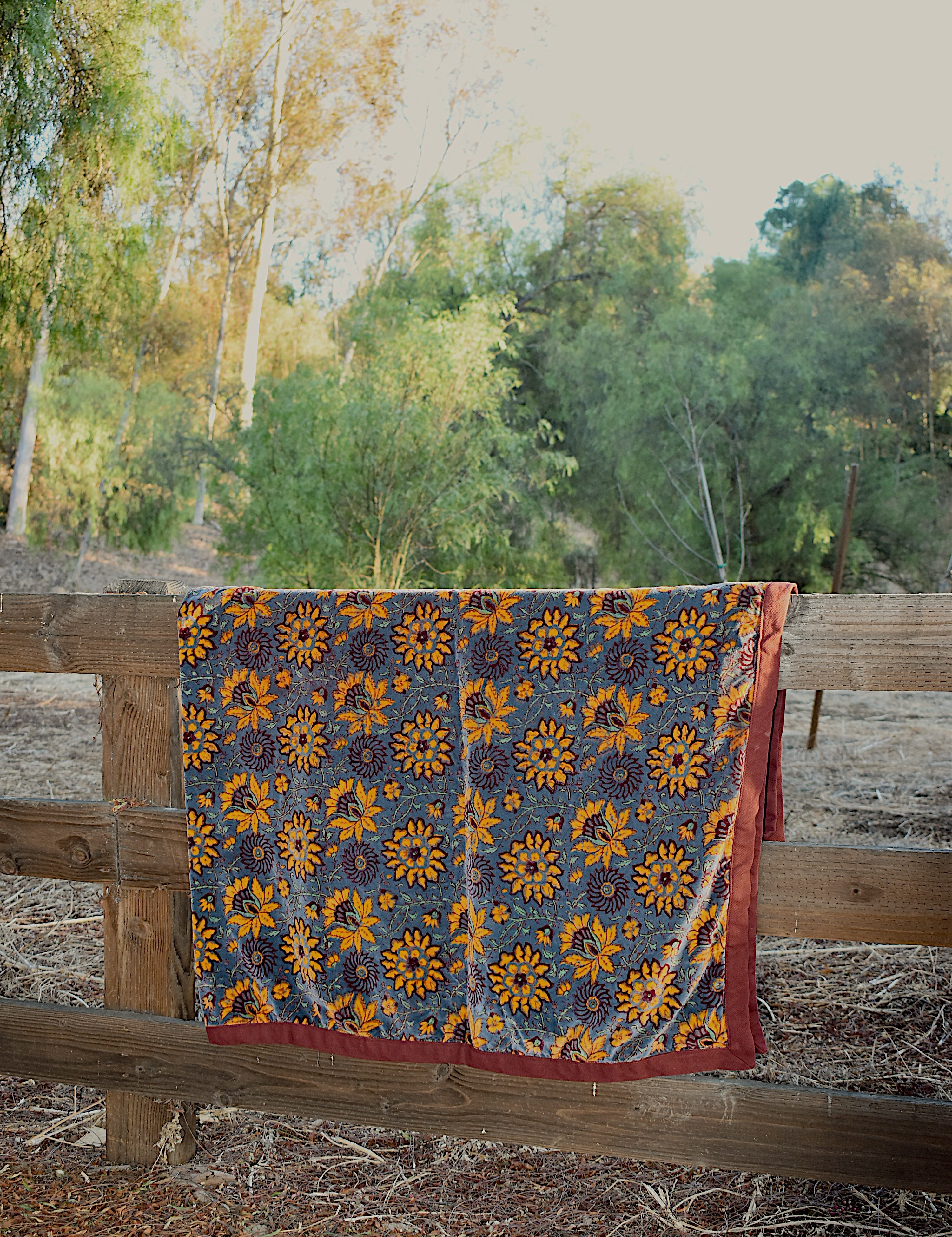 Blue velvet blanket with large yellow floral print and red cotton piping draped over a wooden fence in a rustic setting with trees in the background.