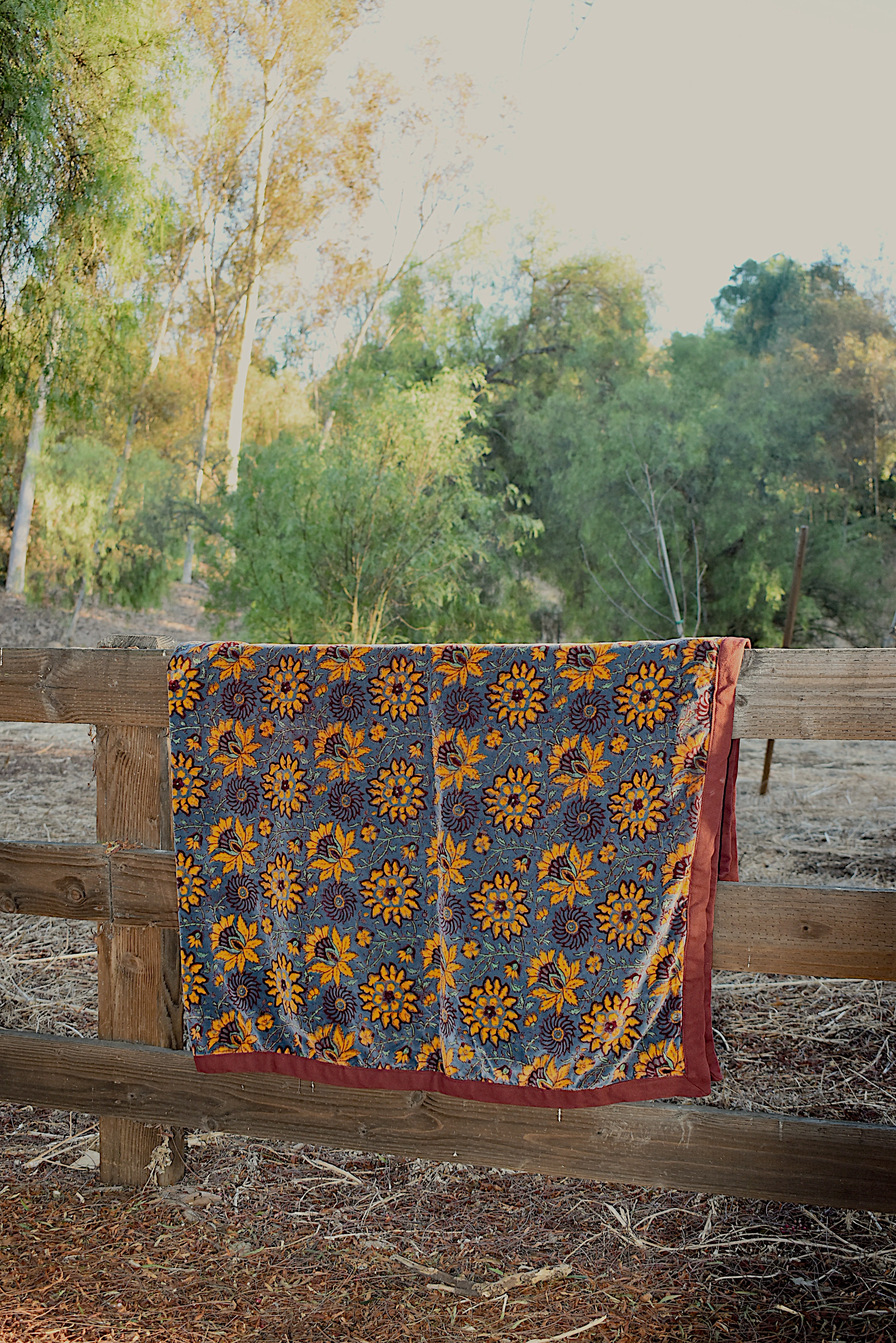 Blue velvet blanket with large yellow floral print and red cotton piping draped over a wooden fence in a rustic setting with trees in the background.
