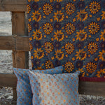 A rustic outdoor scene showing a dark blue velvet blanket with a large yellow floral pattern draped over a wooden fence, with two matching decorative throw pillows in front.