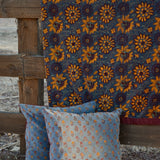 A rustic outdoor scene showing a dark blue velvet blanket with a large yellow floral pattern draped over a wooden fence, with two matching decorative throw pillows in front.