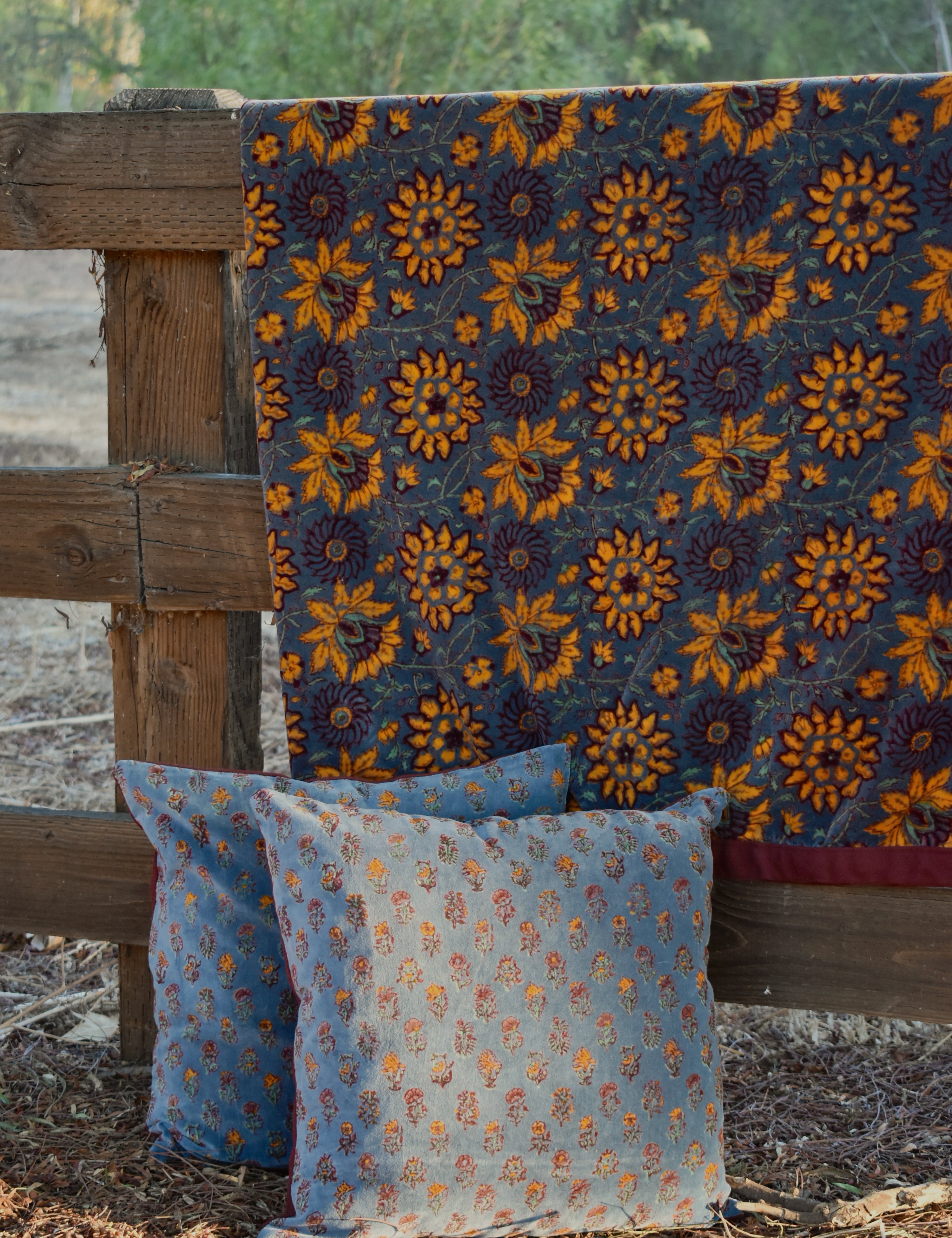 A rustic outdoor scene showing a dark blue velvet blanket with a large yellow floral pattern draped over a wooden fence, with two matching decorative throw pillows in front.