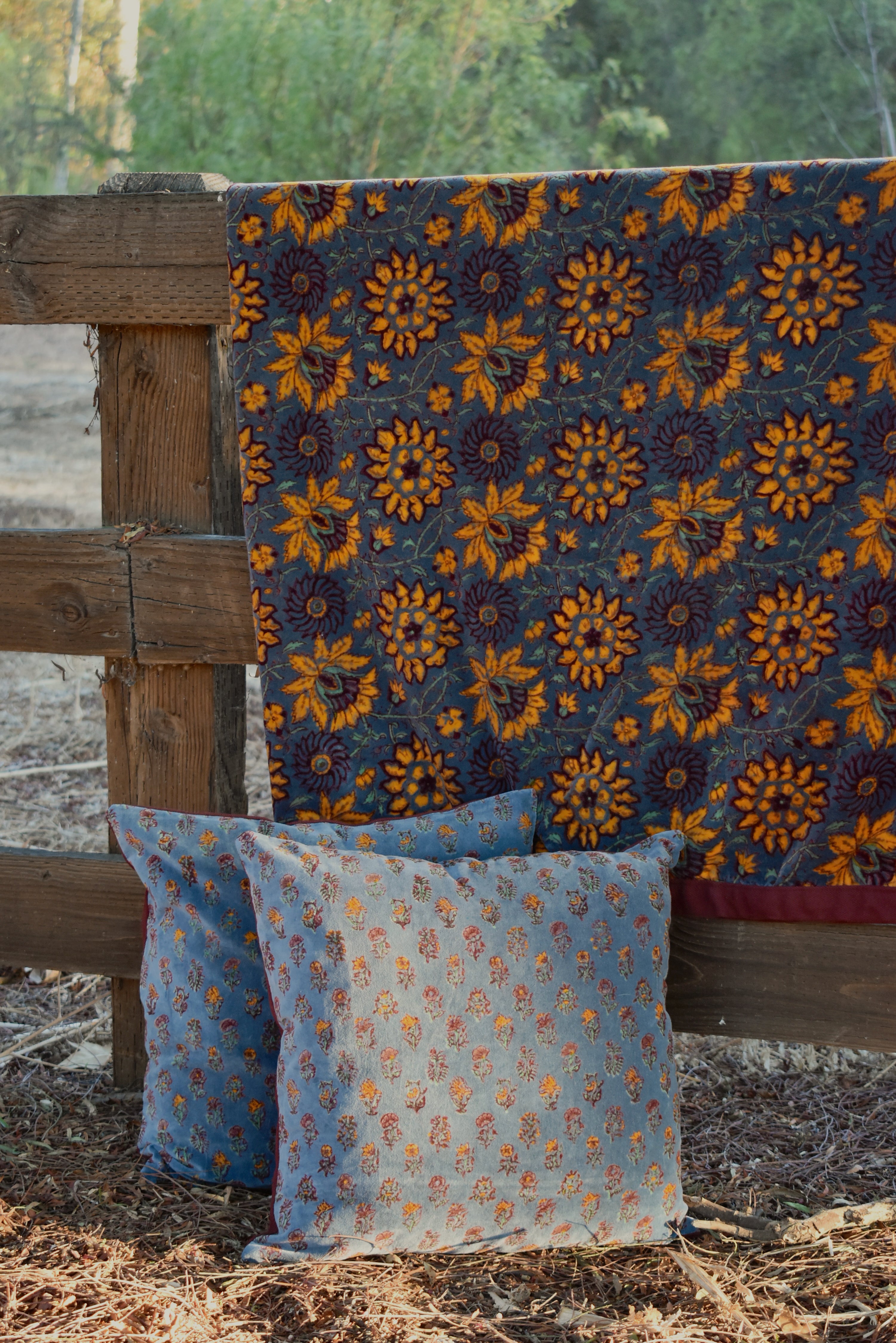 A rustic outdoor scene showing a dark blue velvet blanket with a large yellow floral pattern draped over a wooden fence, with two matching decorative throw pillows in front.