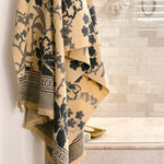 A beige cotton bath towel with a dark navy floral pattern hangs from a hook in a tiled bathroom.