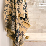 A beige cotton bath towel with a dark navy floral pattern hangs from a hook in a tiled bathroom.