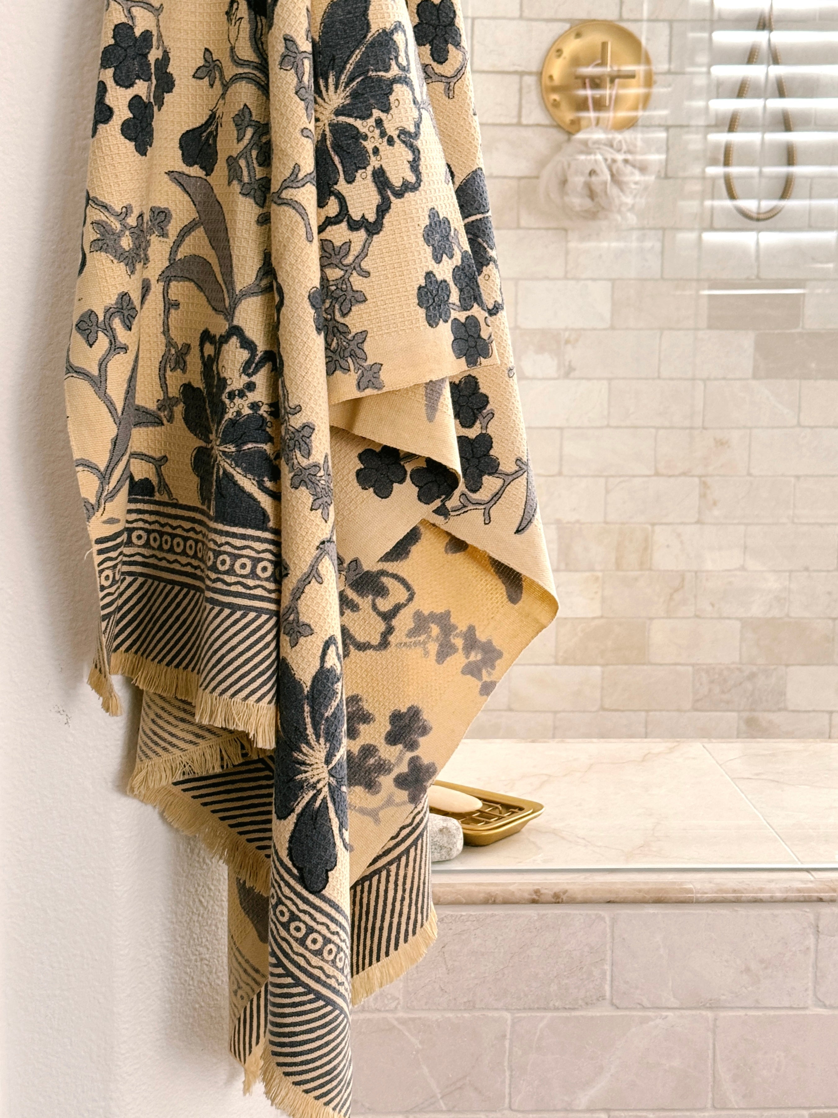 A beige cotton bath towel with a dark navy floral pattern hangs from a hook in a tiled bathroom.