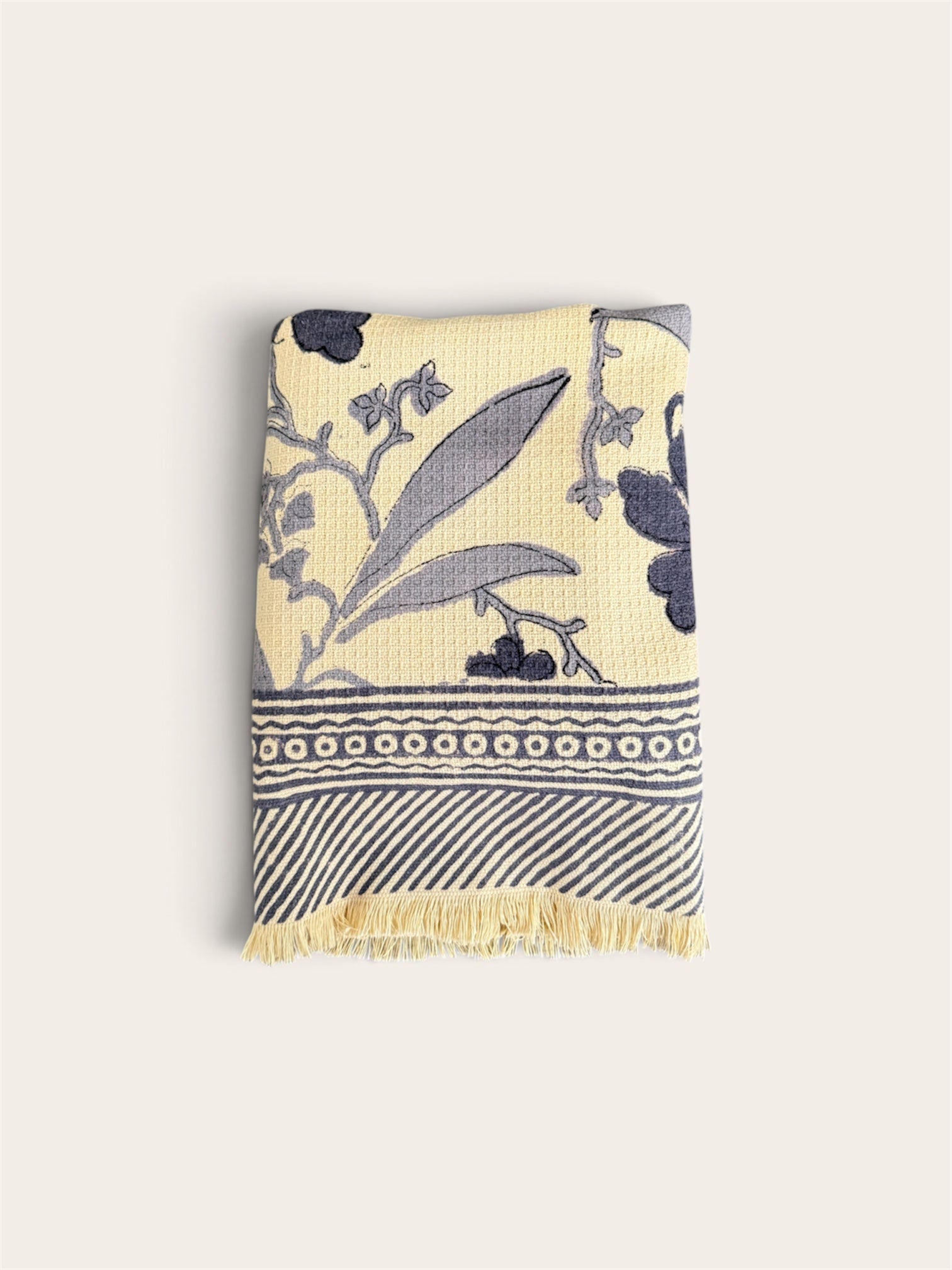 A folded waffle weave bath towel with a cream background and navy blue botanical and geometric patterns, featuring a fringed edge, shown against a solid off-white background.