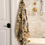 A beige bath towel with a dark blue floral pattern and buttercream fringe, hanging from a brass hook on a white wall in a modern bathroom with beige tile and gold fixtures.