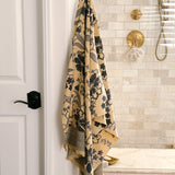 A beige bath towel with a dark blue floral pattern and buttercream fringe, hanging from a brass hook on a white wall in a modern bathroom with beige tile and gold fixtures.
