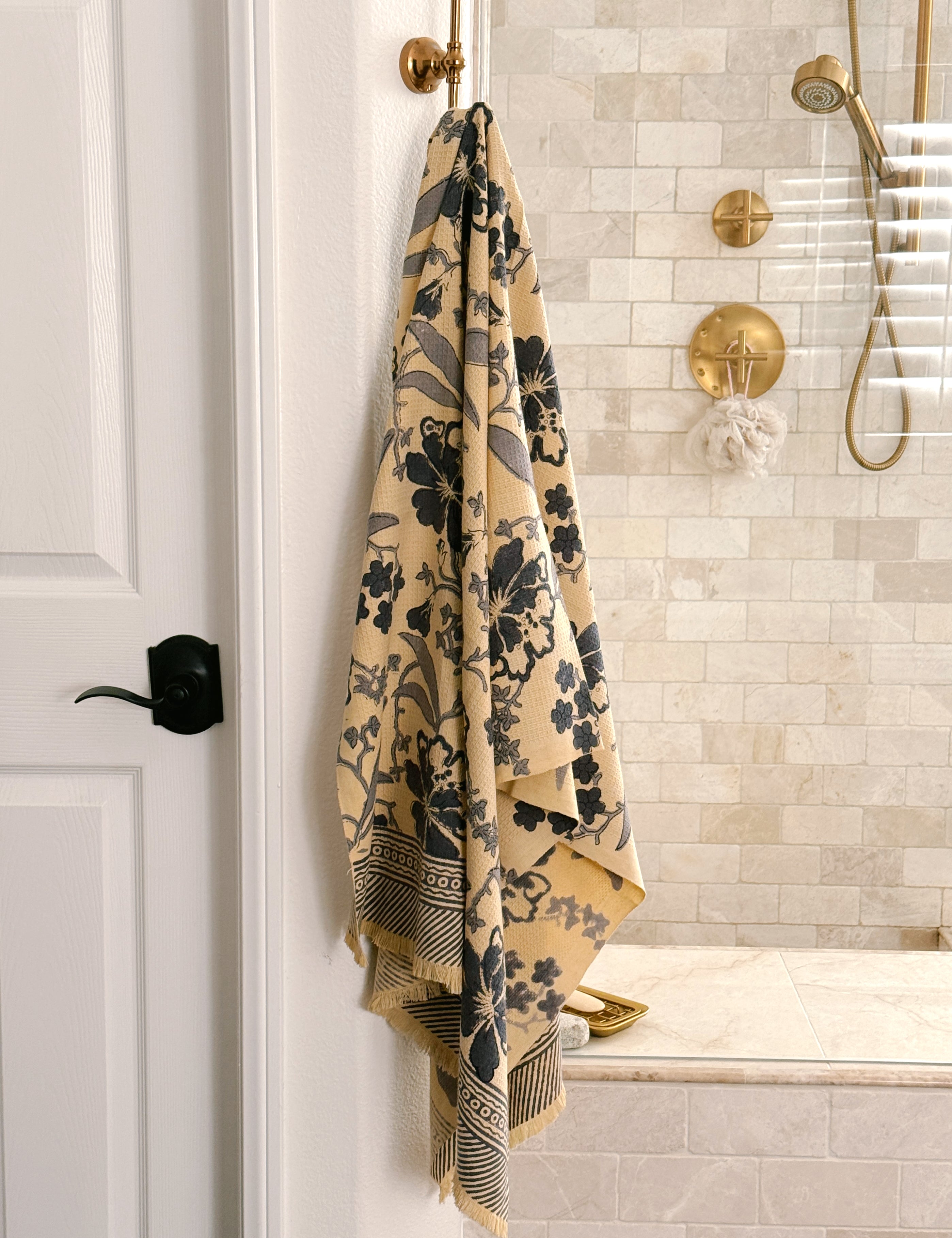 A beige bath towel with a dark blue floral pattern and buttercream fringe, hanging from a brass hook on a white wall in a modern bathroom with beige tile and gold fixtures.