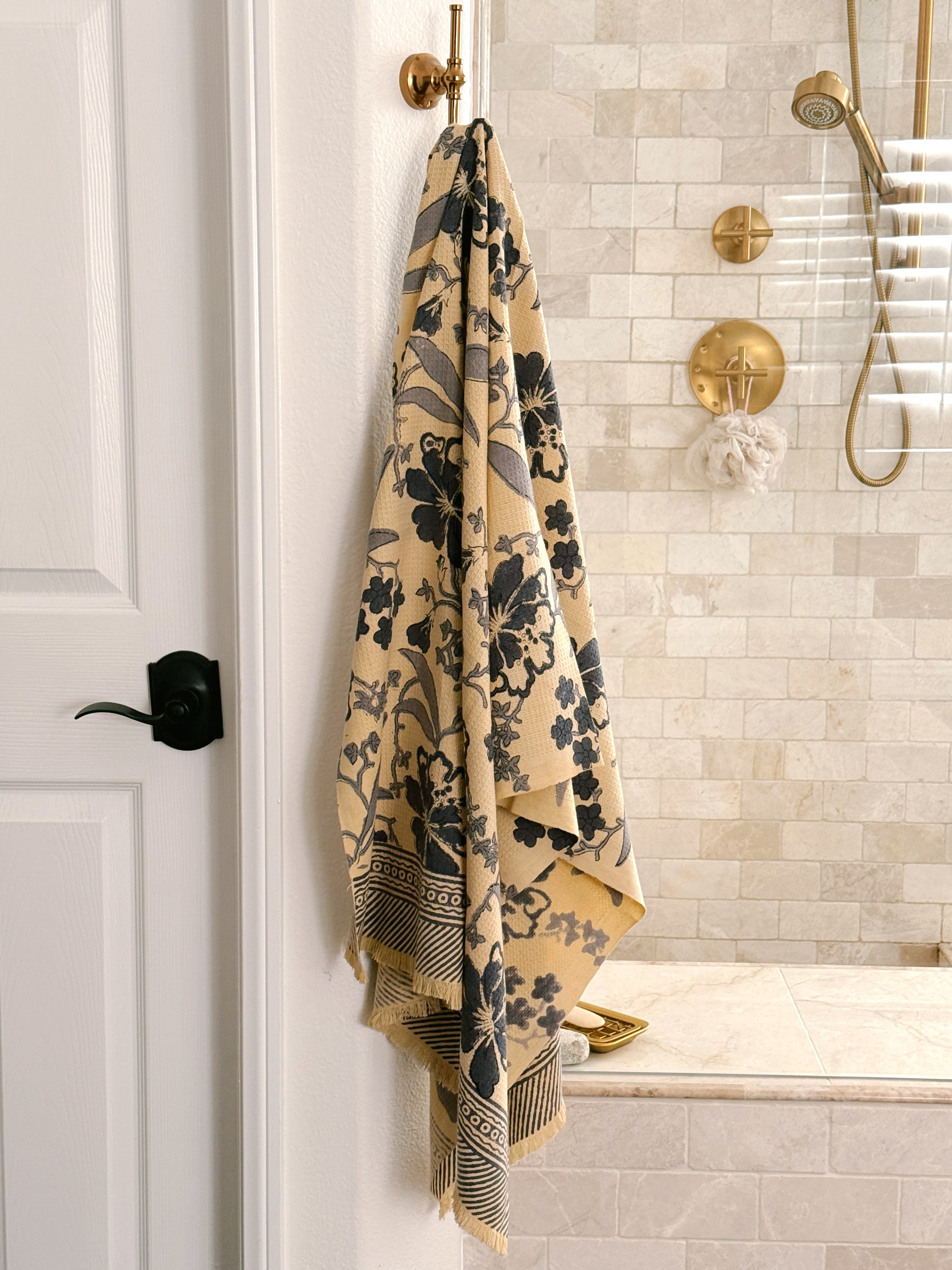 A beige bath towel with a dark blue floral pattern and buttercream fringe, hanging from a brass hook on a white wall in a modern bathroom with beige tile and gold fixtures.