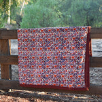 Red and purple floral printed blanket draped over a wooden fence in a rustic setting with trees in the background. 