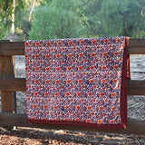 Red and purple floral printed blanket draped over a wooden fence in a rustic setting with trees in the background. 