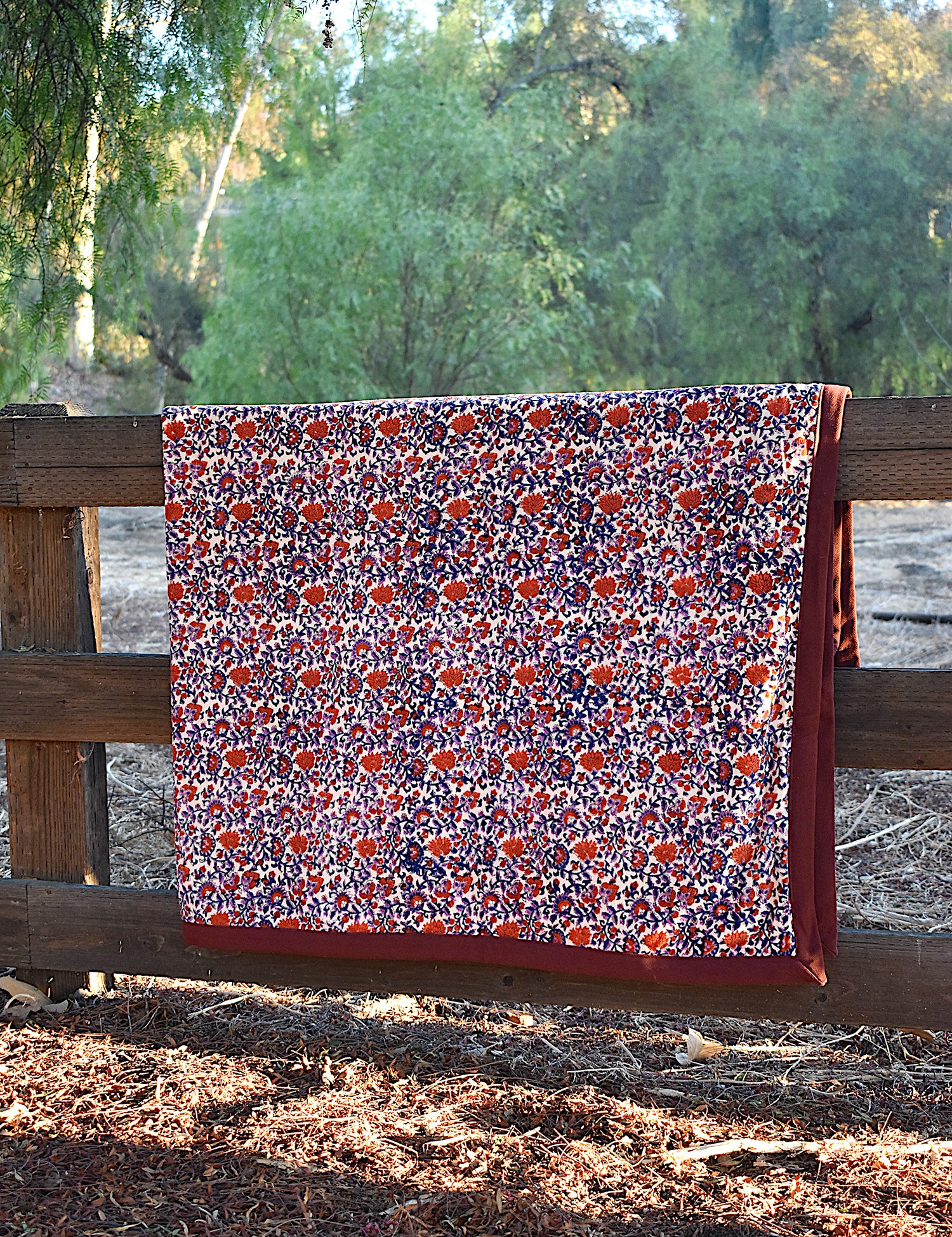 Red and purple floral printed blanket draped over a wooden fence in a rustic setting with trees in the background. 
