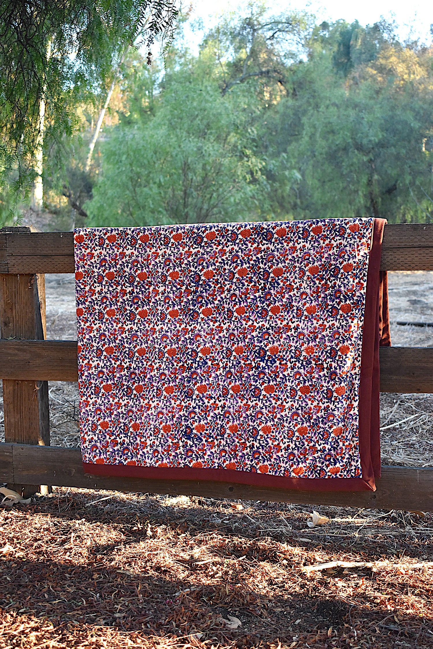 Red and purple floral printed blanket draped over a wooden fence in a rustic setting with trees in the background. 