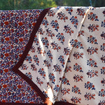 Red and purple floral printed blanket draped over a wooden fence in a rustic setting. Corner of blanket is lifted up to show reversible floral print. 