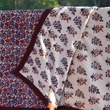 Red and purple floral printed blanket draped over a wooden fence in a rustic setting. Corner of blanket is lifted up to show reversible floral print. 