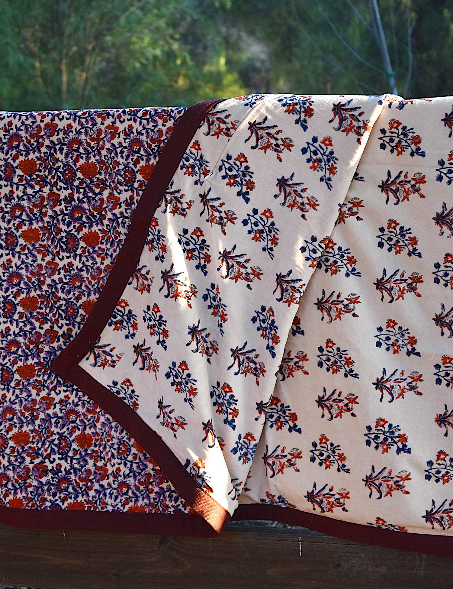 Red and purple floral printed blanket draped over a wooden fence in a rustic setting. Corner of blanket is lifted up to show reversible floral print. 