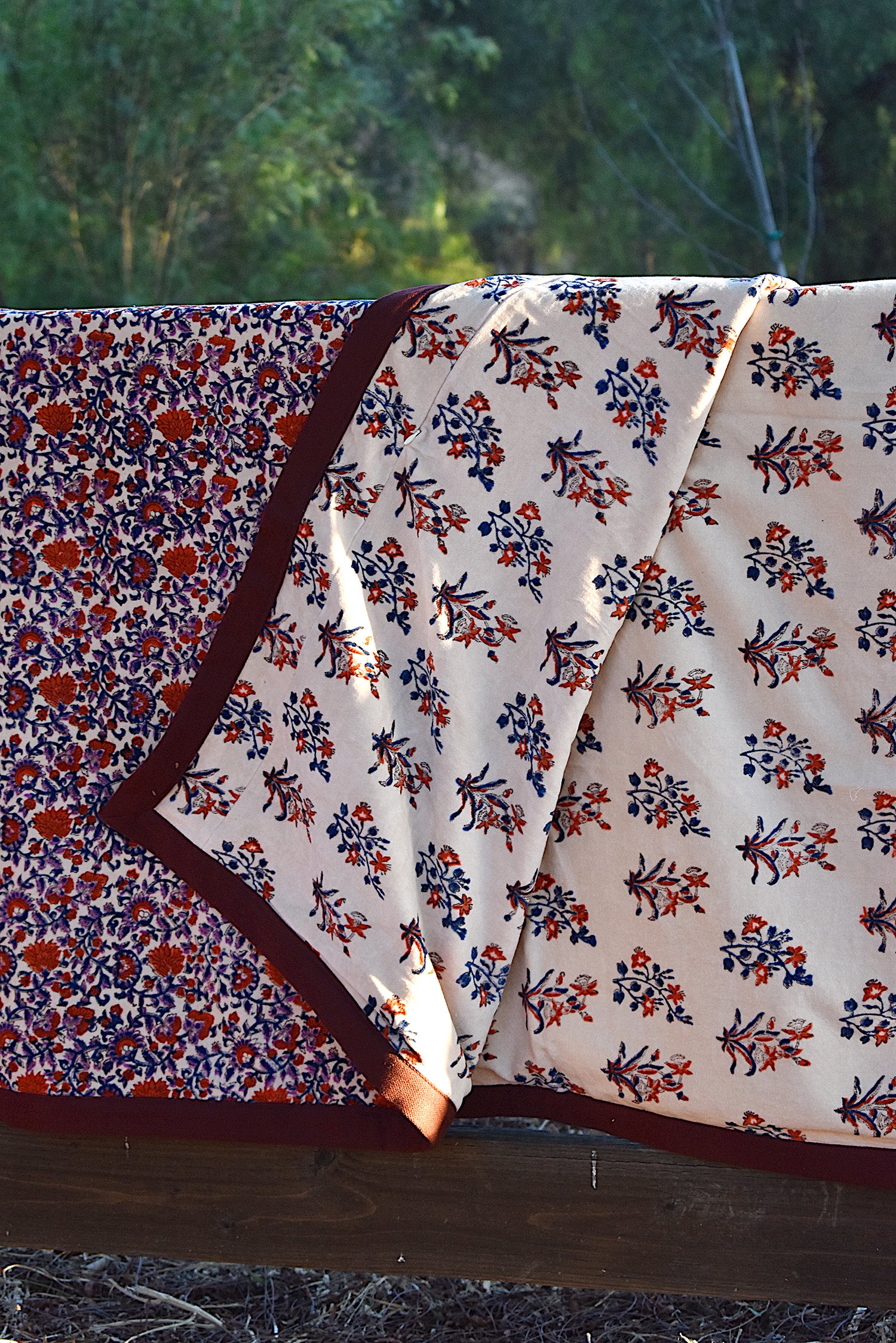 Red and purple floral printed blanket draped over a wooden fence in a rustic setting. Corner of blanket is lifted up to show reversible floral print. 