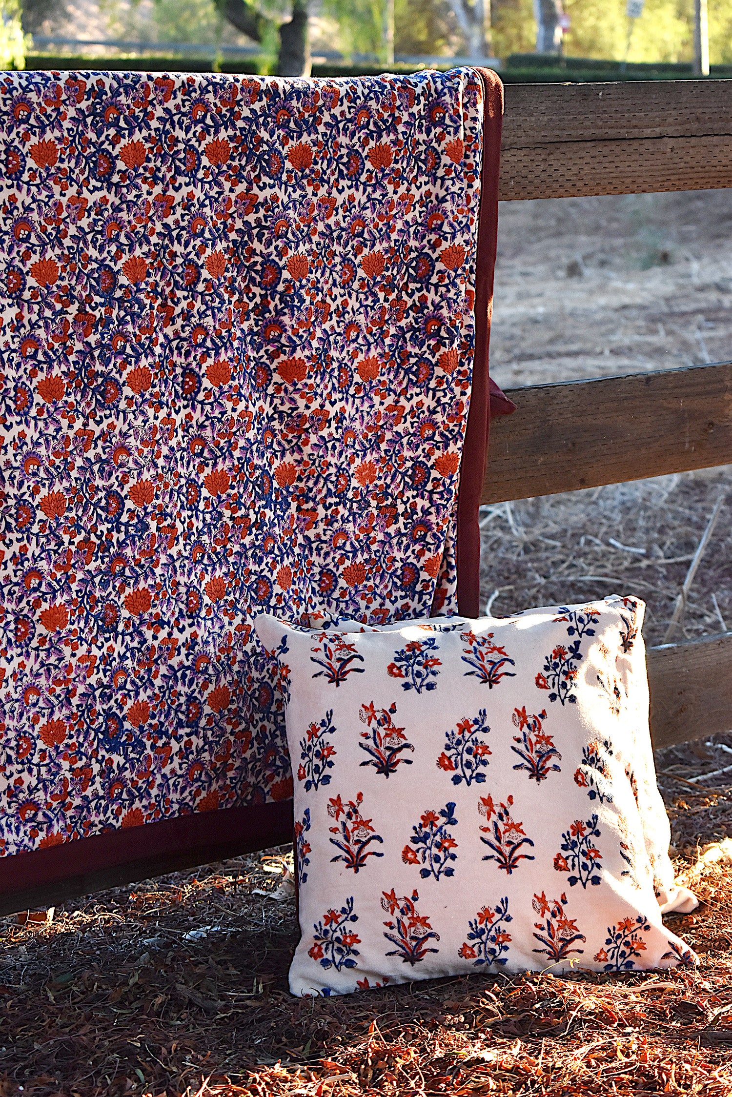 Blanket with red and purple floral pattern draped over a wooden fence in rustic setting. Two pillows with floral print on the ground in front of blanket. 