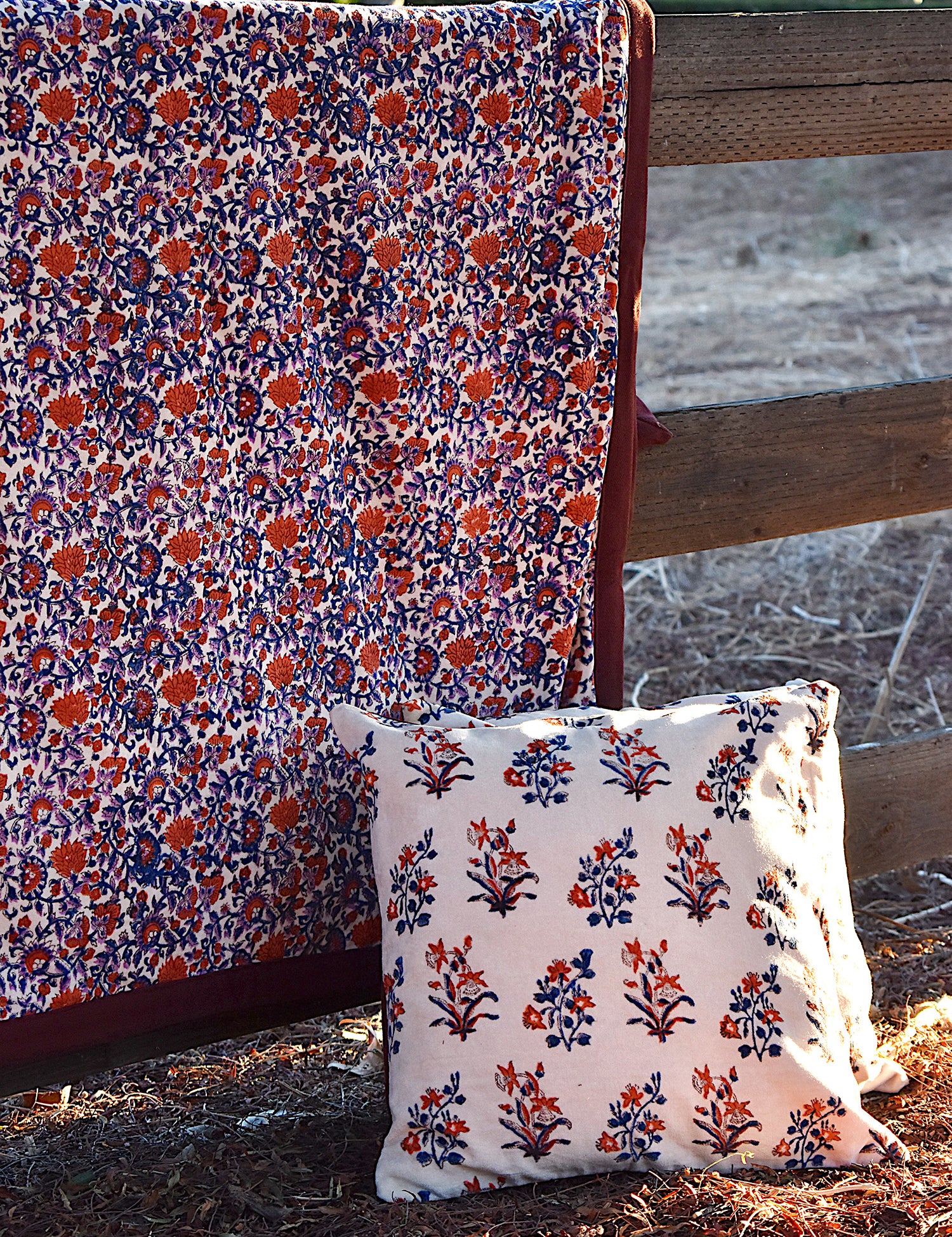 Floral printed blanket draped over a wooden fence in a rustic setting. Floral printed pillows sitting on ground in front of blanket.