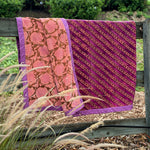 A folded patterned velvet blanket, draped over a wooden fence outdoors. The fabric has two main designs: one pink section with a brown floral vine pattern and purple border, and one magenta-purple section with diagonal gold patterned floral stripes and a purple border.