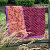 A folded patterned velvet blanket, draped over a wooden fence outdoors. The fabric has two main designs: one pink section with a brown floral vine pattern and purple border, and one magenta-purple section with diagonal gold patterned floral stripes and a purple border.