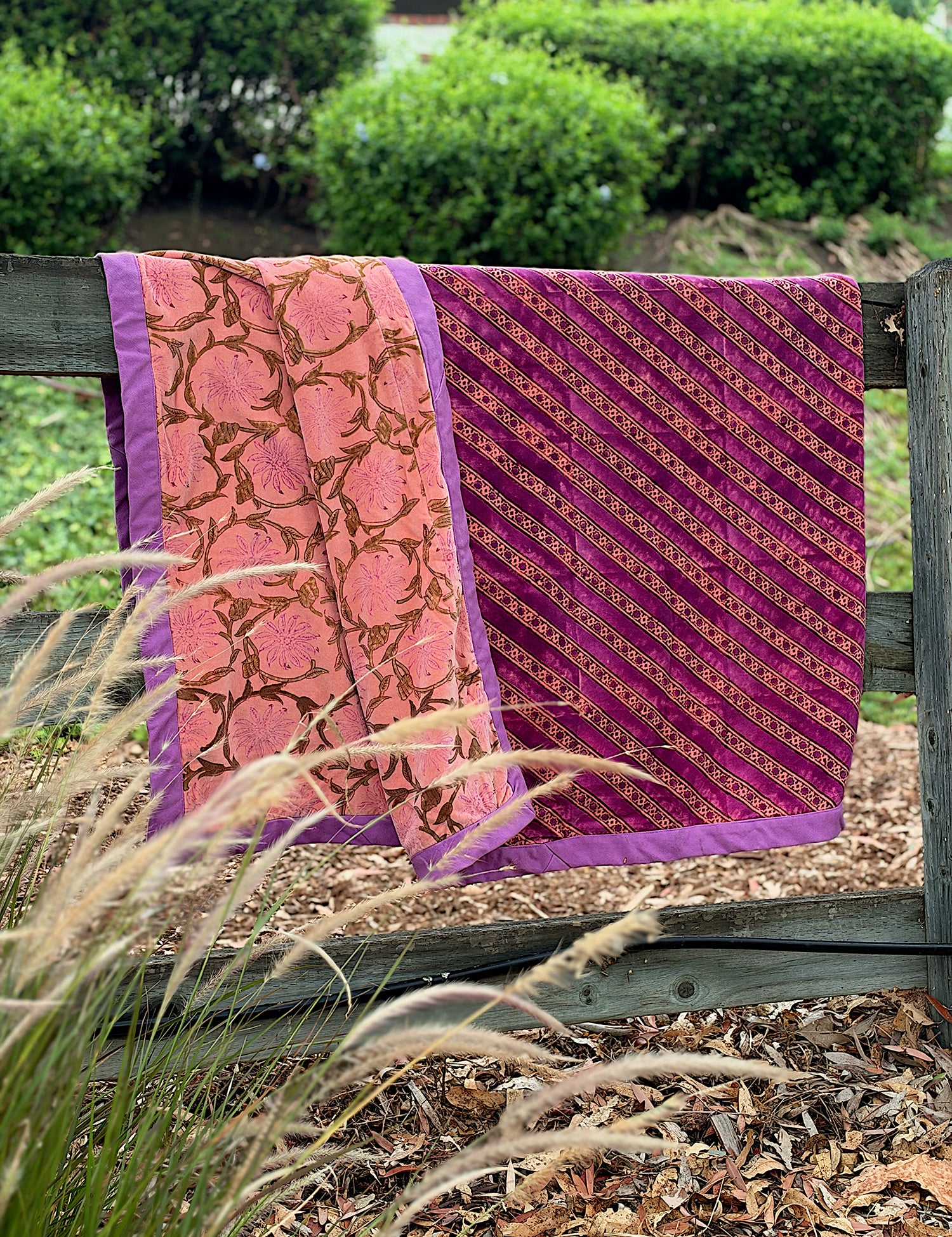 A folded patterned velvet blanket, draped over a wooden fence outdoors. The fabric has two main designs: one pink section with a brown floral vine pattern and purple border, and one magenta-purple section with diagonal gold patterned floral stripes and a purple border.