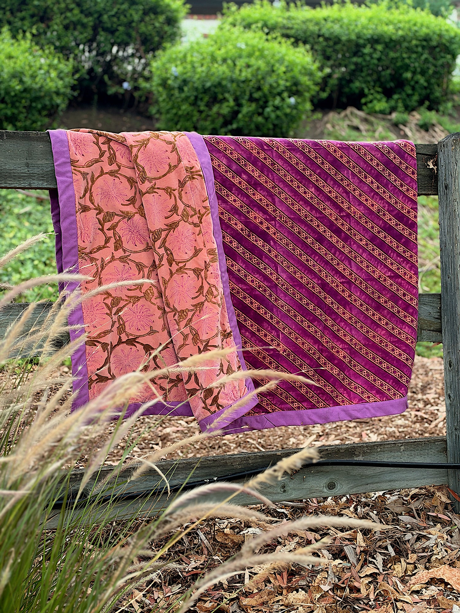 A folded patterned velvet blanket, draped over a wooden fence outdoors. The fabric has two main designs: one pink section with a brown floral vine pattern and purple border, and one magenta-purple section with diagonal gold patterned floral stripes and a purple border.