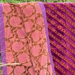 A folded and draped velvet blanket, featuring a pink floral pattern on one side and a diagonal gold floral pattern on the other, shown outdoors against a foliage background.
