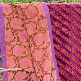 A folded and draped velvet blanket, featuring a pink floral pattern on one side and a diagonal gold floral pattern on the other, shown outdoors against a foliage background.