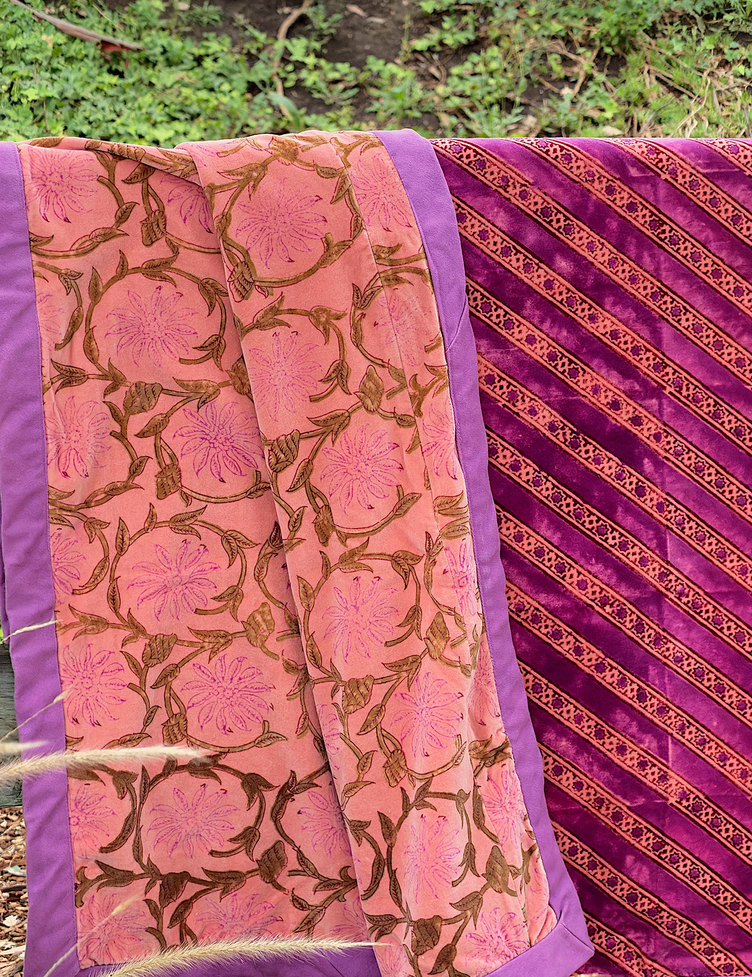 A folded and draped velvet blanket, featuring a pink floral pattern on one side and a diagonal gold floral pattern on the other, shown outdoors against a foliage background.