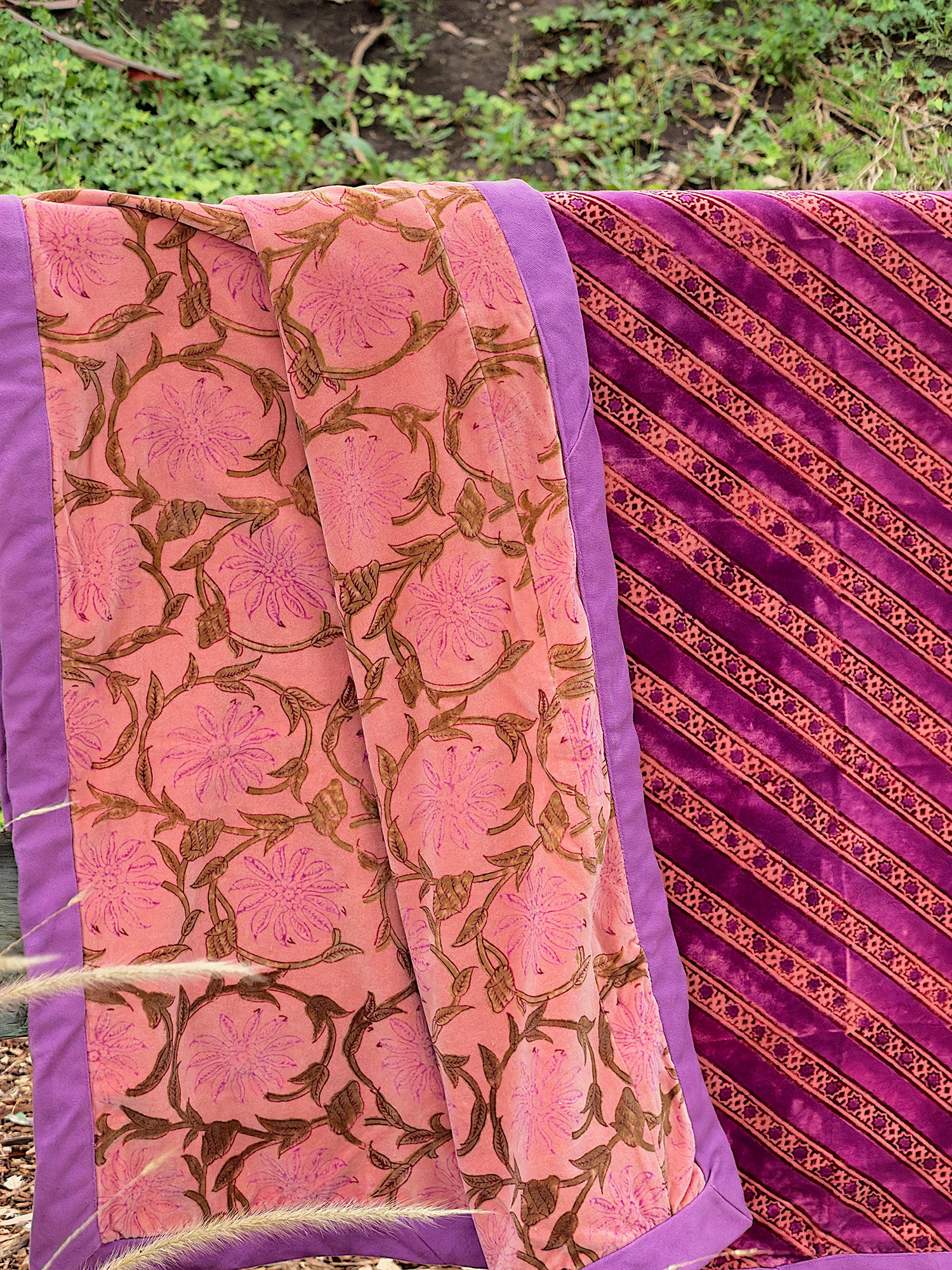 A folded and draped velvet blanket, featuring a pink floral pattern on one side and a diagonal gold floral pattern on the other, shown outdoors against a foliage background.