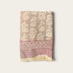 A folded woven textile, a bath towel featuring a beige and cream floral pattern on the upper section, a red and cream geometric border, and a red and cream striped section with cream fringe at the bottom, set against a solid off-white background.