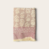 A folded woven textile, a bath towel featuring a beige and cream floral pattern on the upper section, a red and cream geometric border, and a red and cream striped section with cream fringe at the bottom, set against a solid off-white background.